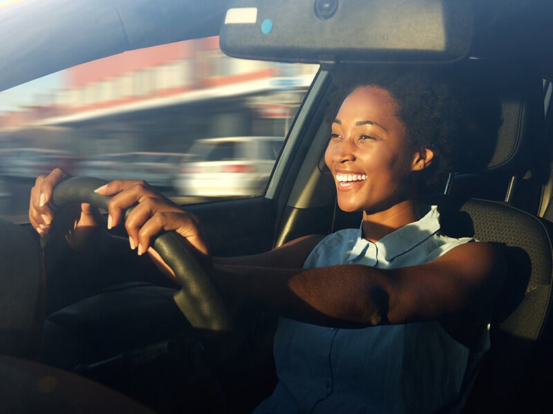 A happy Black woman driving a car with sunlight on her face and blurred motion visible through the window.