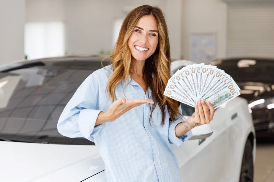 Smiling woman in a light blue shirt holding a fan of hundred-dollar bills while standing next to a white car.