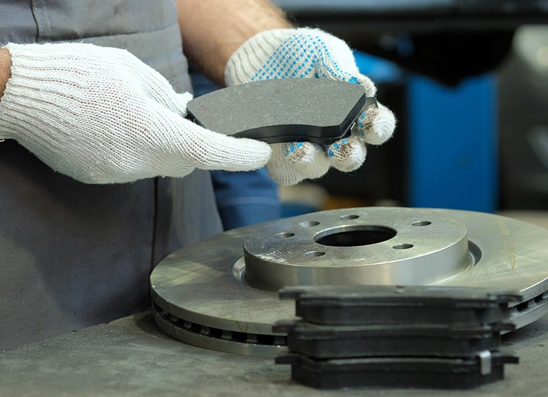 Gloved hands holding new brake pads over a new brake disc and more pads on a work surface.