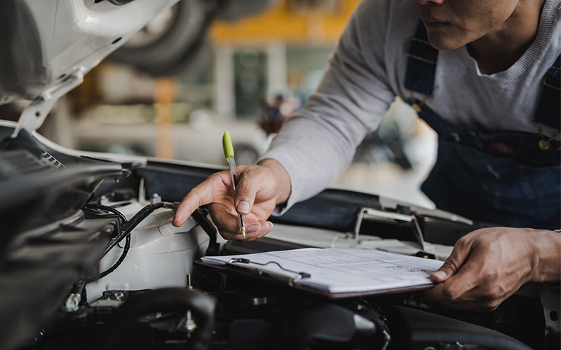 Mechanic in overalls pointing at a car engine while holding a pen and clipboard.