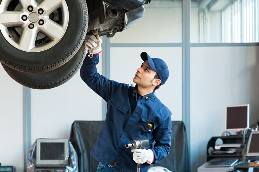 A male auto mechanic in a blue uniform and cap inspects a car's underside, holding an impact wrench.