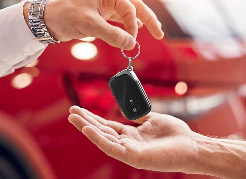 A hand handing a black car remote key on a keyring to another outstretched hand, with a red car in the background.